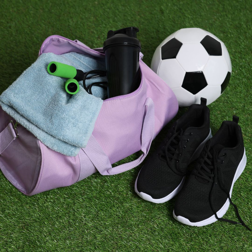 Purple bag with sports equipment, black shoes, and a soccer ball on grass.