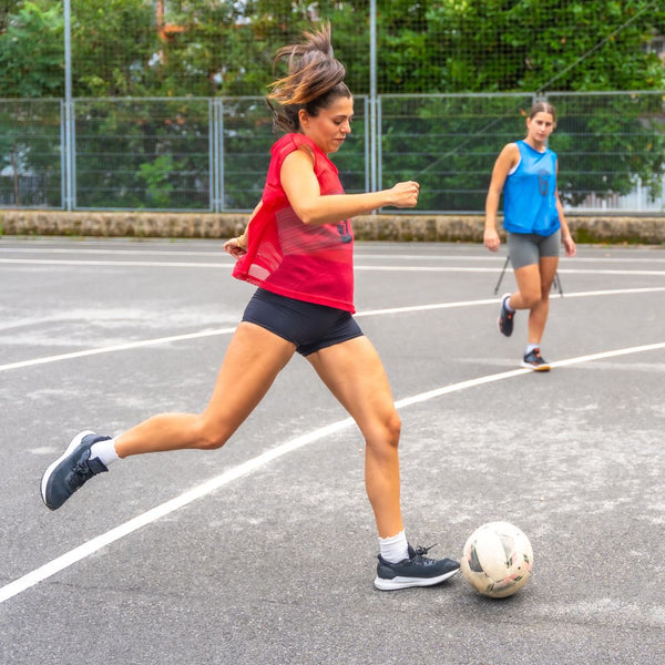 Two women playing soccer on an outdoor court with a fence and trees in the background.